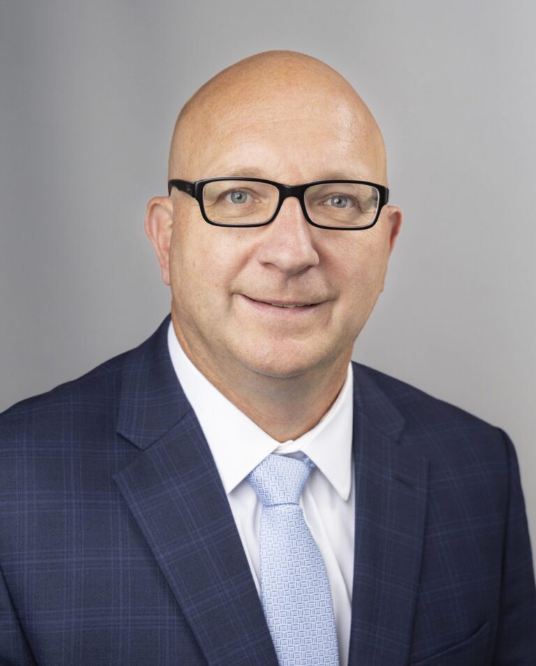 Bald man with glasses wearing a blue checked suit, white shirt, and light blue tie poses for a professional headshot against a plain grey background.