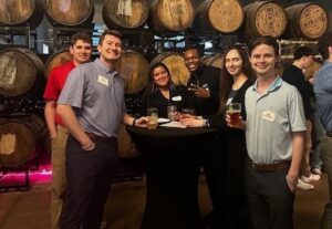 Six people stand around a cocktail table with drinks in hand, smiling, in front of stacked wooden barrels at an indoor event.