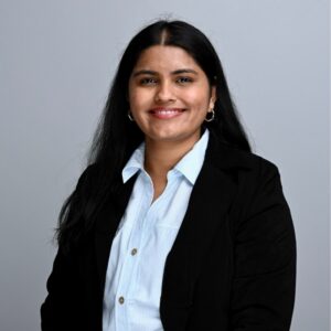 A woman with long black hair wearing a black blazer and light blue shirt smiles in front of a plain gray background.
