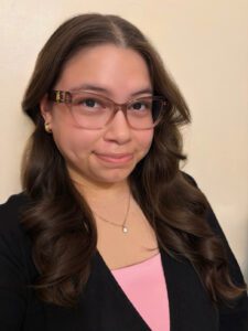 A woman with long brown hair and glasses is wearing a black cardigan over a pink top, smiling slightly in front of a plain light-colored wall.